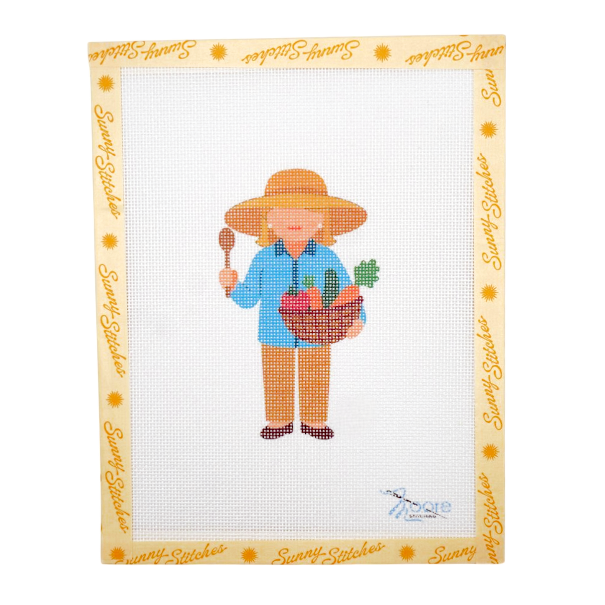 A framed cross-stitch design on white Aida fabric, featuring Martha Stewart in a straw hat holding a basket of vegetables, with a decorative border printed with the brand name 'Sunny Stitches'.
