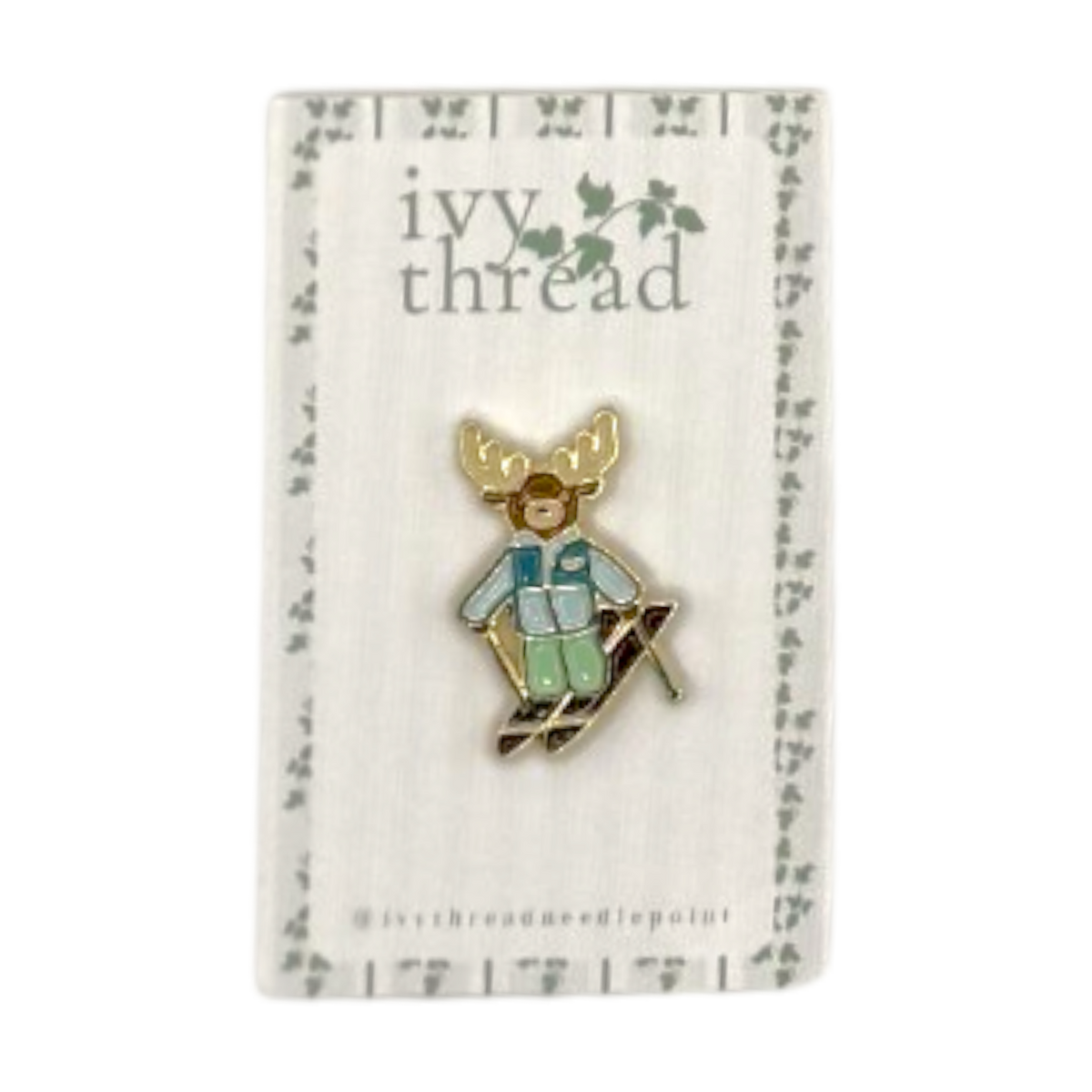 A small enamel pin of a moose skier wearing a light blue jacket and green pants, mounted on a white display card with the brand name 'ivy thread' printed at the top.