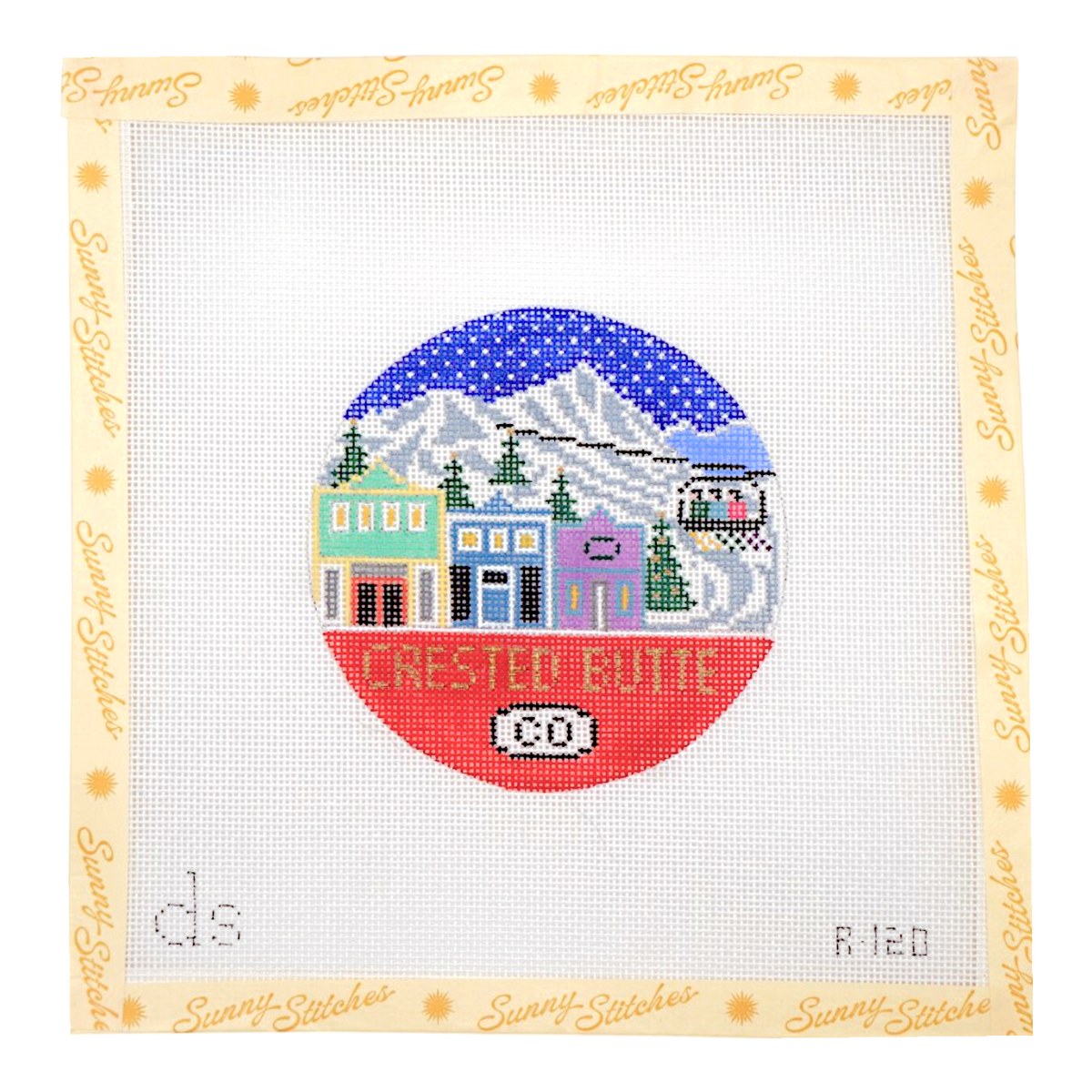 A square cross-stitch pattern on a white grid fabric with a cream border printed with the brand name 'Sunny Stitches' in gold. The central stitched design is a circular winter village scene labeled 'Crested Butte' with a blue starry sky, mountains, colorful buildings, trees, and a red ground section.