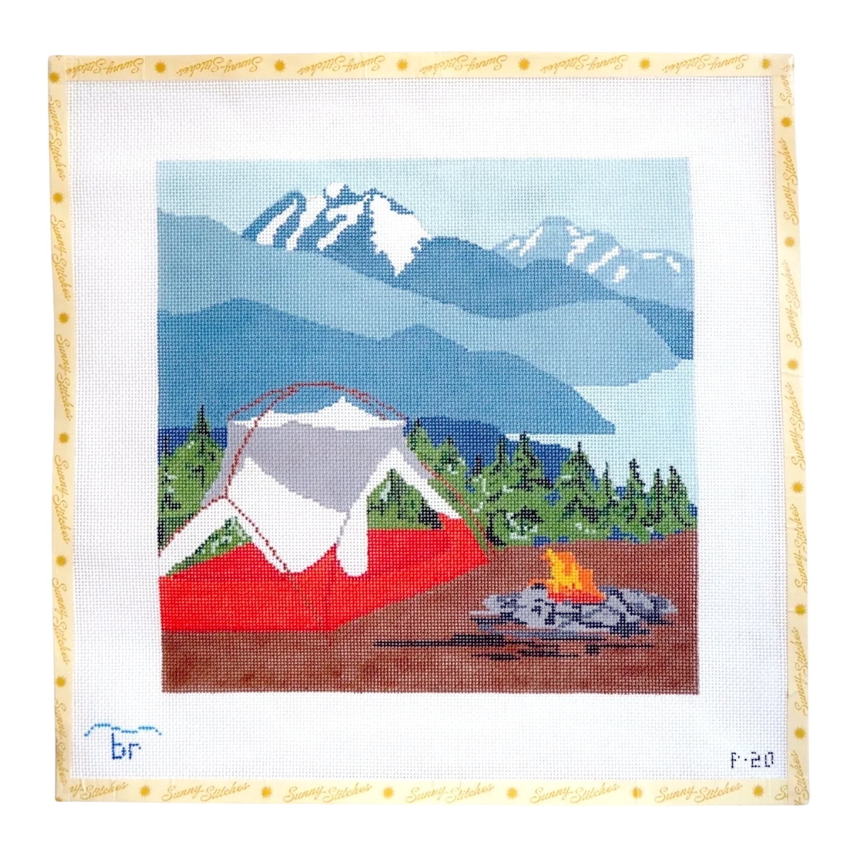 A square needlepoint canvas featuring a mountain landscape with blue and gray peaks, a red tent, green trees, a lake, and a campfire. The canvas has a white border with a decorative gold dotted frame and small handwritten markings in the bottom corners.